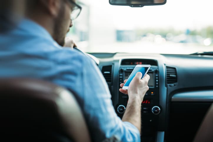 A distracted driver looks at his phone screen and has his other hand on the wheel