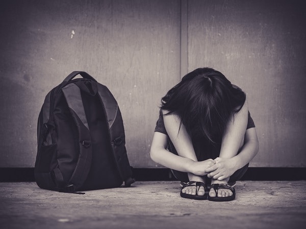 Black and white photo of child with head on knees, sitting next to backpack