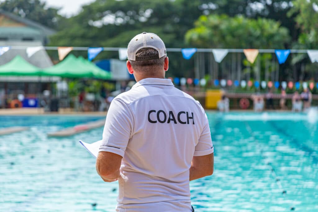 Image of swimming coach facing toward the pool with shirt that says “coach”
